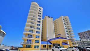 Um hotel com vários andares, varandas, um toldo azul e uma área de estacionamento com carros.
