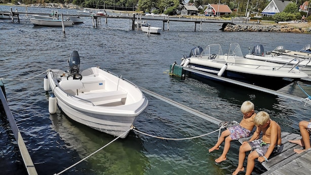 Une jetée en bois avec un bateau amarré, une vue dégagée sur l’eau et une côte lointaine parsemée de bâtiments.