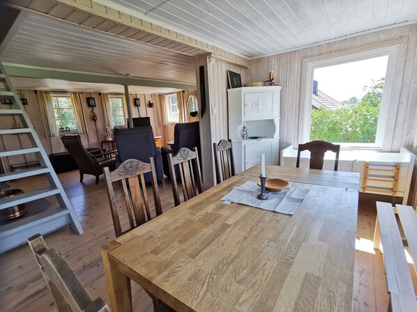 A cozy room with a wooden floor, a dining table with chairs, a ladder, and a window with a view of greenery.