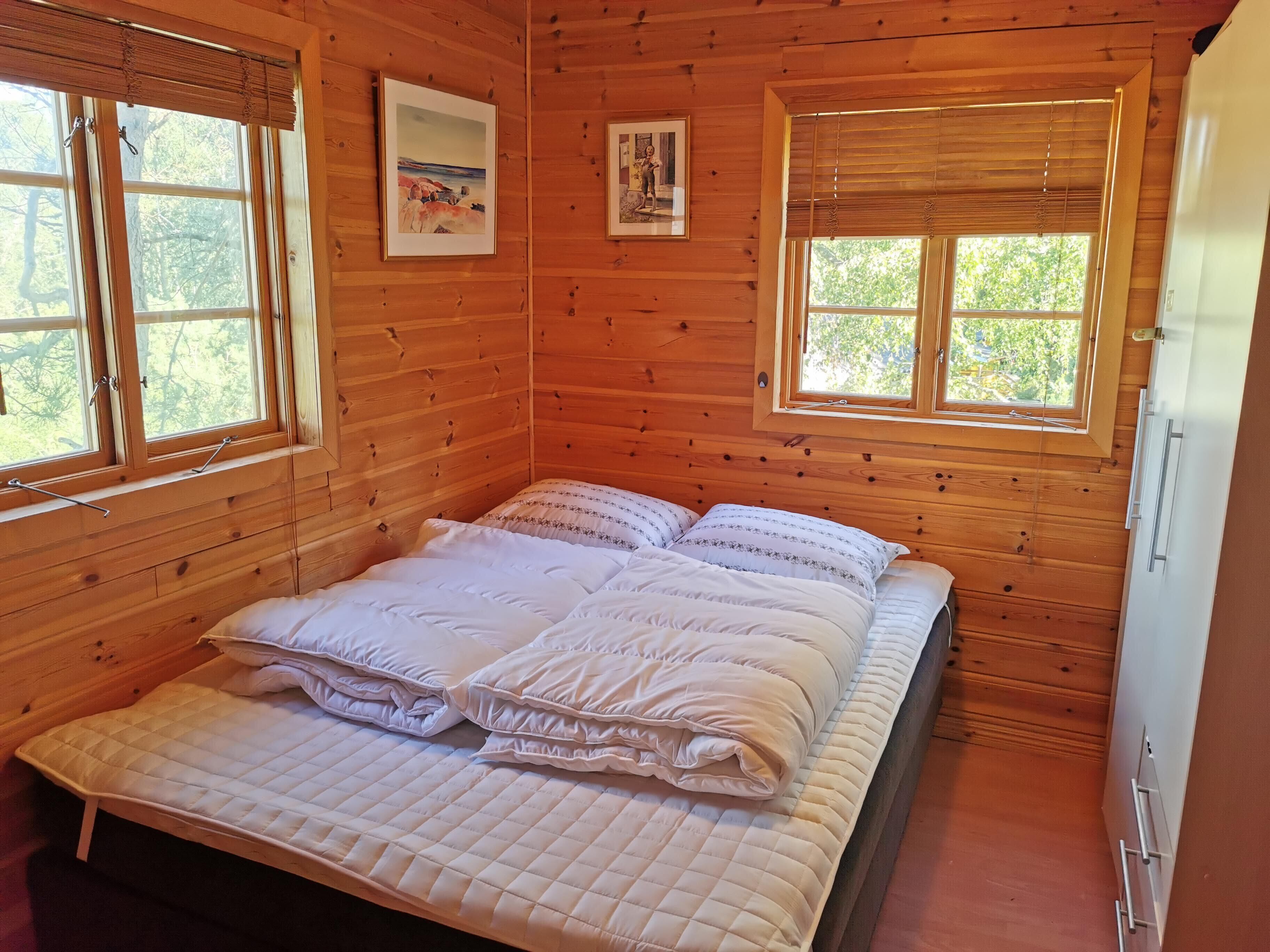 A wooden cabin bedroom with a bed, two windows, and a refrigerator.