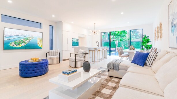 A modern living room with a sofa, ottoman, and coffee table, a kitchen area with bar stools, and large windows offering a view of greenery.