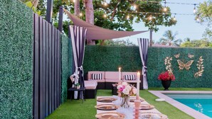 A neatly arranged outdoor dining area with a table set for six, surrounded by a green hedge and a pool.