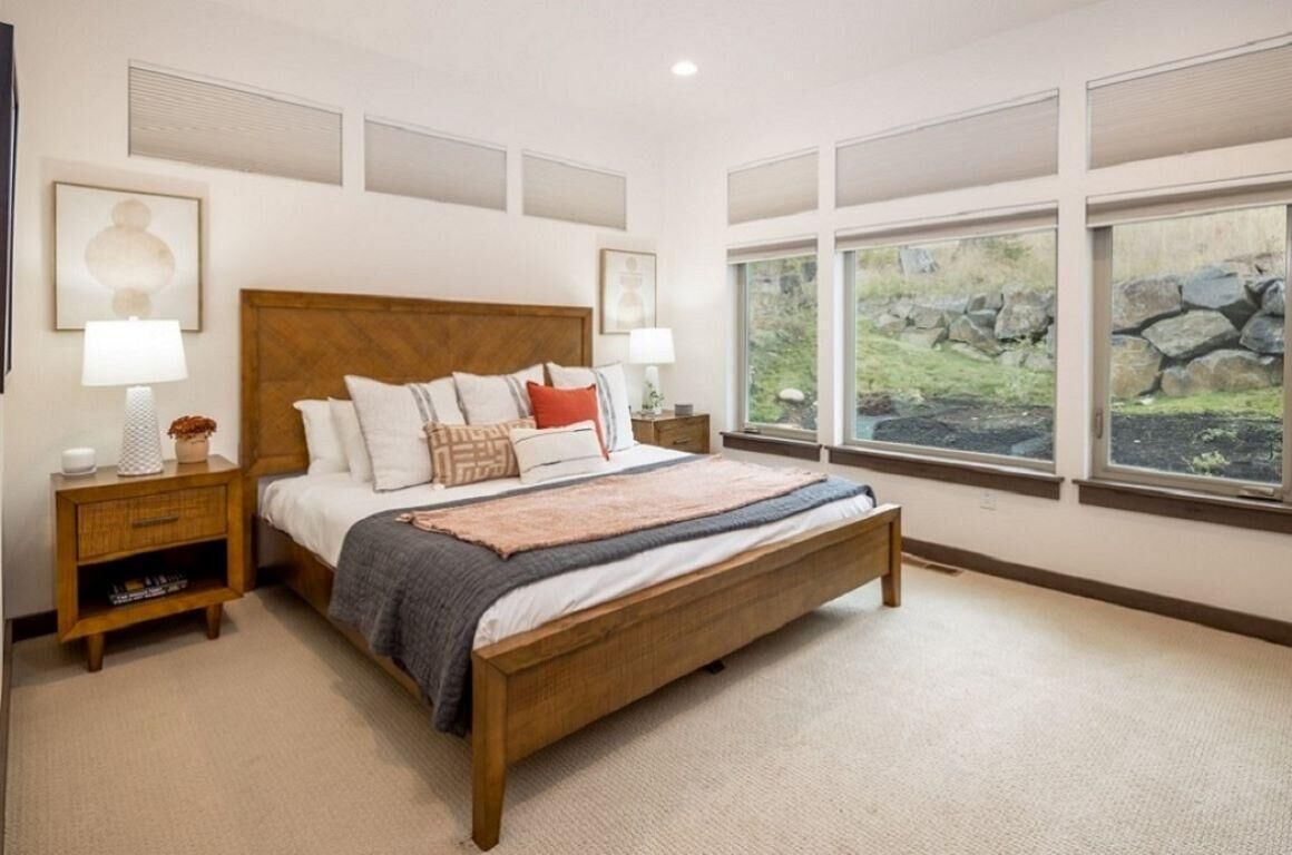 Un dormitorio con cama de madera, dos mesitas de noche, un ventanal con vistas a rocas y vegetación, y obras de arte en las paredes.