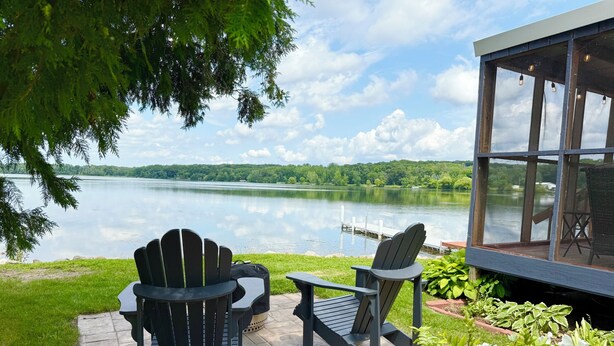 An outdoor seating area with Adirondack chairs and a table overlooking a lake, with a dock and a covered structure in the background.