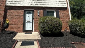 A two-story brick house with a front door, two windows, and a small front yard with mulch and a bush.