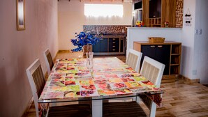 Comedor con una mesa de vidrio, sillas blancas y un mantel colorido. En el centro, un jarrón con flores azules.