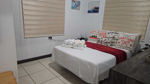 Hotellihuoneessa on valkoisilla lakanoilla peitetty sänky, punainen peitto ja useita koristetyynyjä, joissa on loma-aiheisia kuvioita. Huoneessa on pieni pöytä, jonka päällä on valkoinen pöytäliina ja muutamia esineitä. Huoneessa on ikkuna, jossa on sälekaihtimet, seinään kiinnitetty lämmitin ja ovi, jossa on kahva.