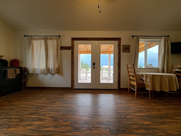 A room with a wooden floor, a table with a tablecloth, a chair, a sofa, a TV, and a view of the outdoors through a door.