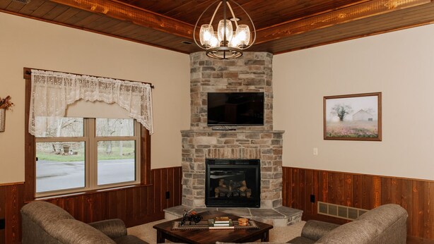 A cozy living room with a stone fireplace, wooden ceiling, and a large window with lace curtains. - Wildflower Paradise: Luxury Lodge Suite in Amish Country (Dundee)