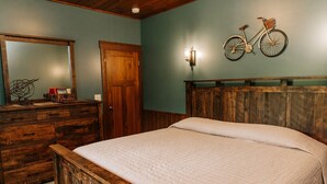 A bedroom with a wooden headboard, a bed with a quilt, a dresser with drawers, a mirror, a wall-mounted bicycle, and a wall sconce. - Adventure Awaits Suite: Luxury, Lodge Suite in Amish Country, Ohio. (Dundee)