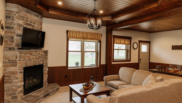 A cozy living room with a stone fireplace, wooden ceiling, and a television mounted above it. There are two windows with curtains, a sofa, and a coffee table with decorative items. - Adventure Awaits Suite: Luxury, Lodge Suite in Amish Country, Ohio. (Dundee)