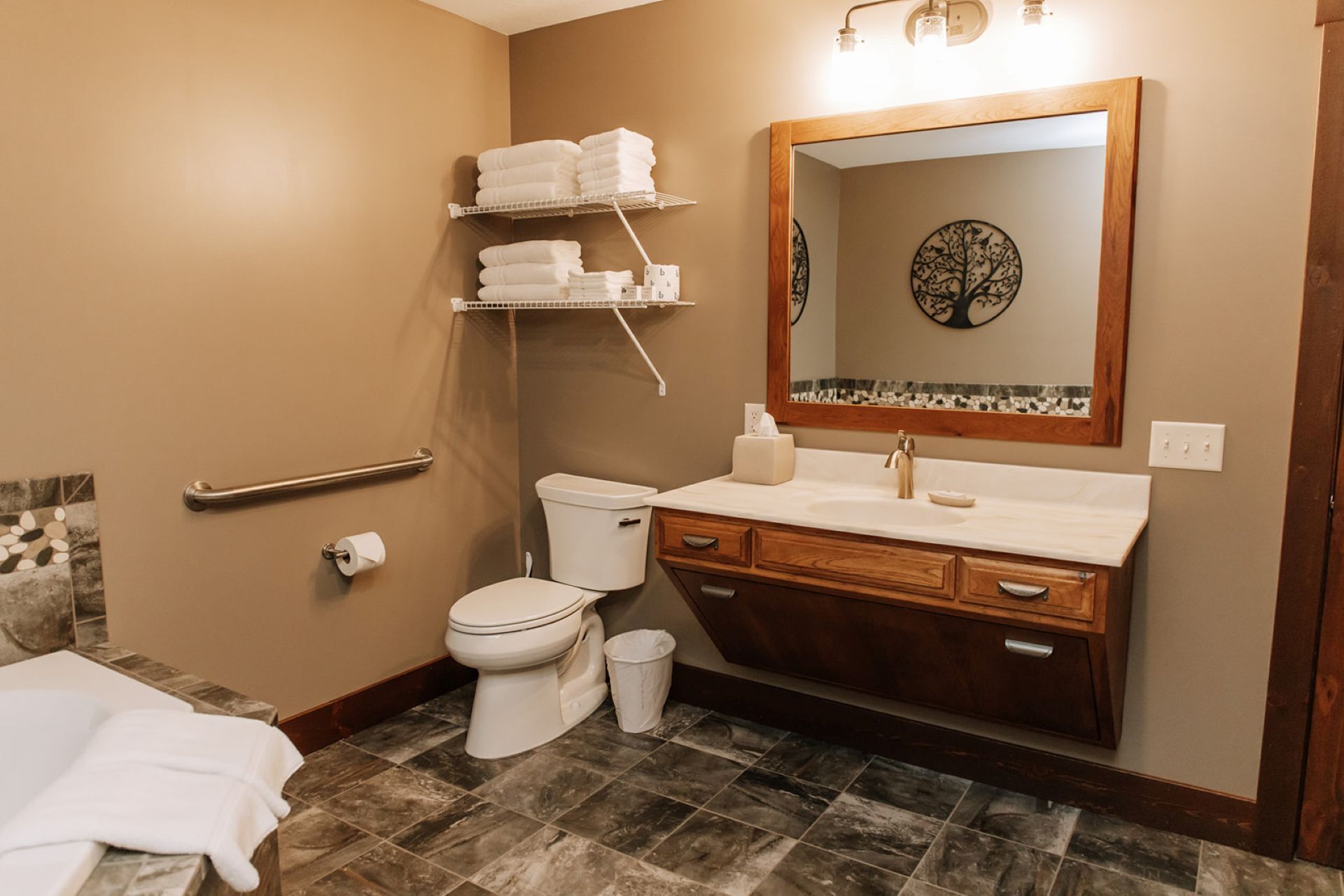 Ein Badezimmer mit Toilette, Waschbecken und Spiegel. Handtücher liegen auf einem Regal, eine Haltestange ist an der Wand angebracht.