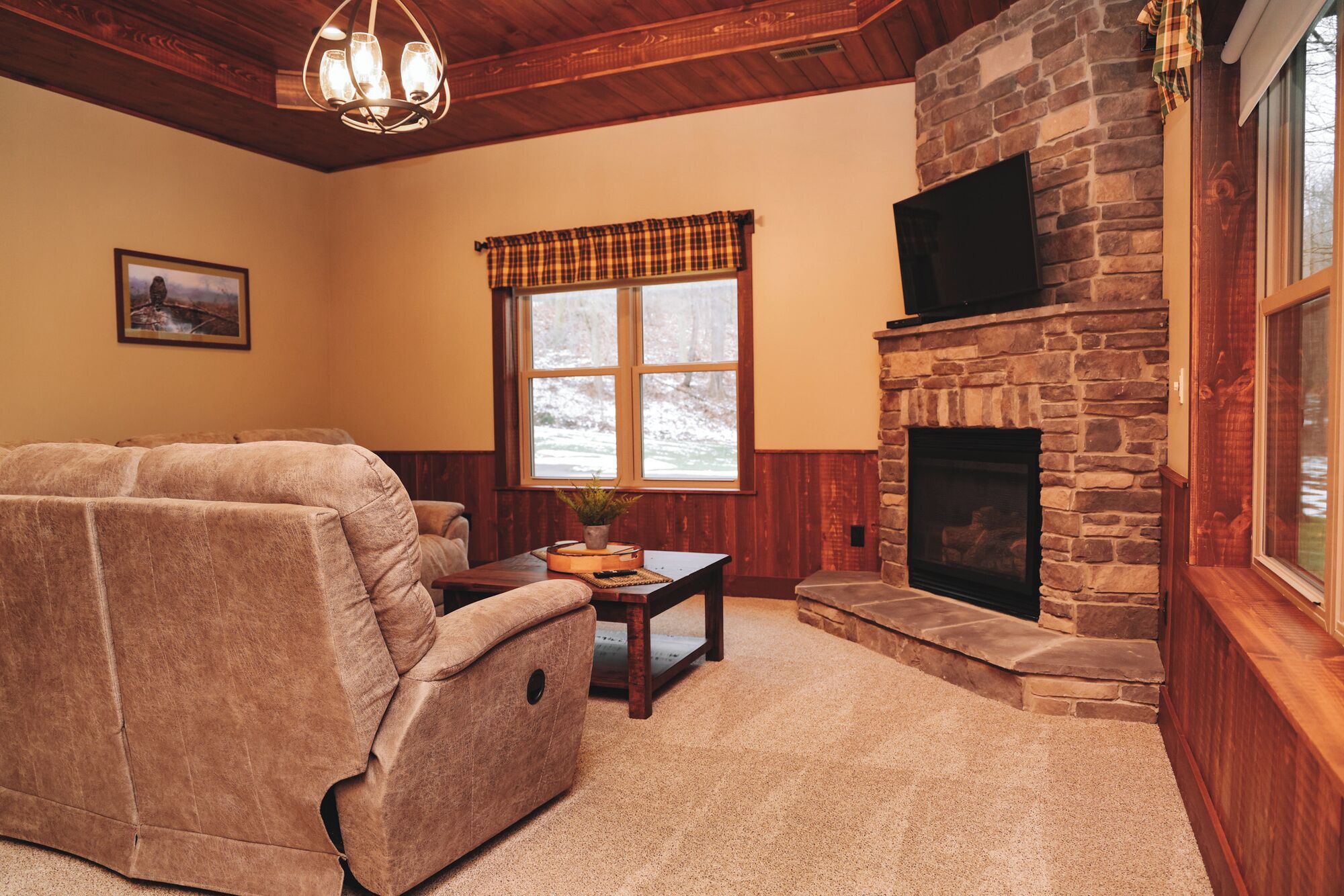 A cozy living room with a stone fireplace, a large window with plaid curtains, and a reclining sofa.