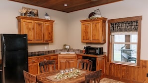 Una cocina con armarios de madera, un refrigerador negro, una mesa de comedor con sillas, un microondas y una ventana con cortinas de pata de gallo.