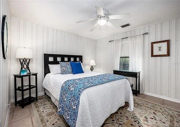 Un dormitorio ordenado con una cama, dos mesitas de noche, un ventilador de techo y una ventana con cortinas.