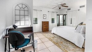 Un dormitorio con cama, escritorio con silla, ventilador de techo y ventana con cortinas.