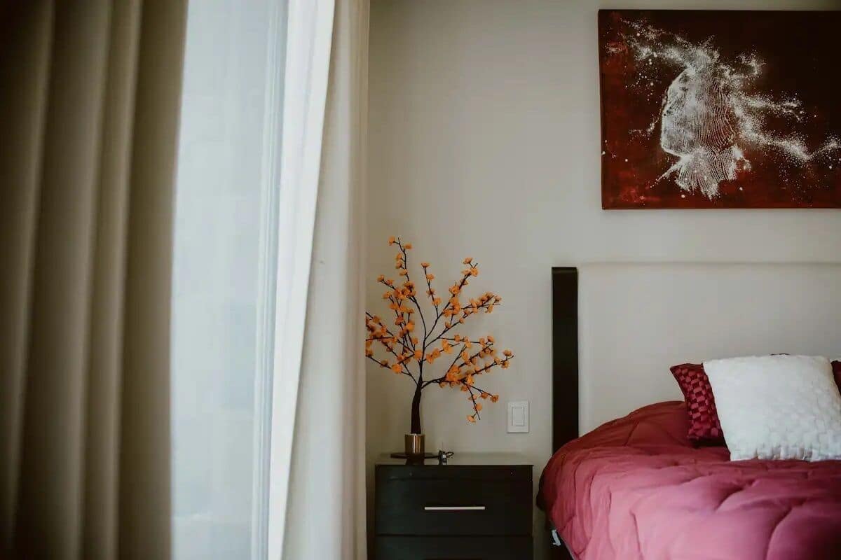 Ein Schlafzimmer mit einem Bett, einem Nachttisch, einer Vase mit Zweigen und einem Bild an der Wand.