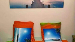 A bed with two pillows featuring ocean scenes, a green blanket, and a painting of a pier on the wall. - Large secure studio, 50 meters from Bas du Fort beach. (Le Gosier)