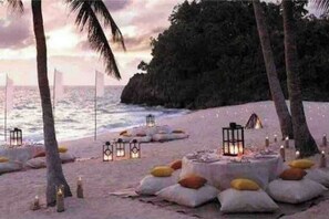 A beach setting with lanterns and cushions arranged on the sand, palm trees, and a view of the ocean. - Large secure studio, 50 meters from Bas du Fort beach. (Le Gosier)