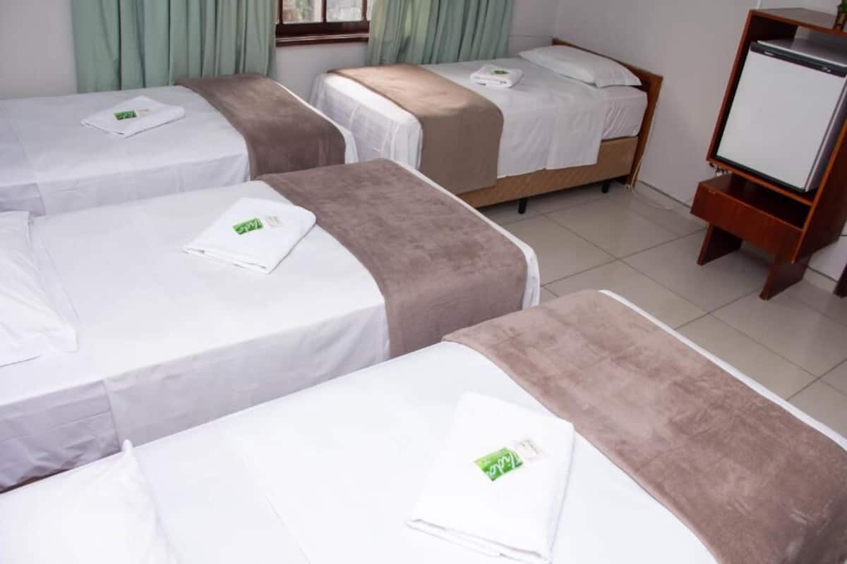 A hotel room with four single beds, each with a white bedspread and a brown blanket. There is a wooden TV stand with a flat-screen TV on the right side of the room.
