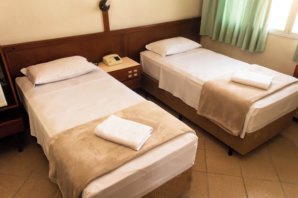 A hotel room with two single beds, each with a pillow and a folded towel. There is a bedside table with a phone and a lamp.