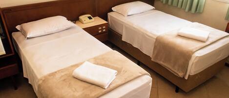 A hotel room with two single beds, each with a pillow and a folded towel. There is a bedside table with a phone and a lamp.