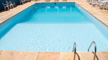 Uma piscina retangular com água azul cristalina, rodeada por espreguiçadeiras brancas e um deck revestido com azulejos.