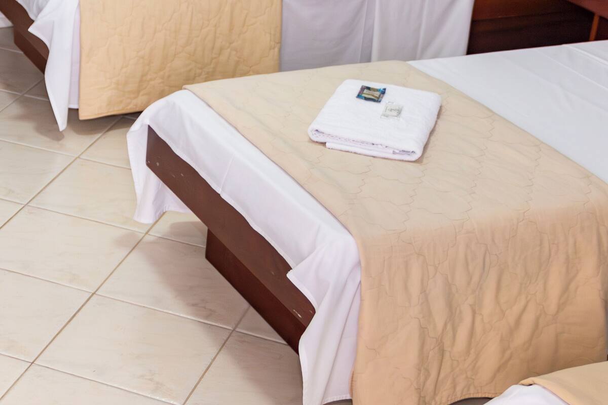 A hotel room with two single beds, each with a beige bedspread and white sheets. There is a wooden headboard on the right bed, and a wooden nightstand with a lamp.