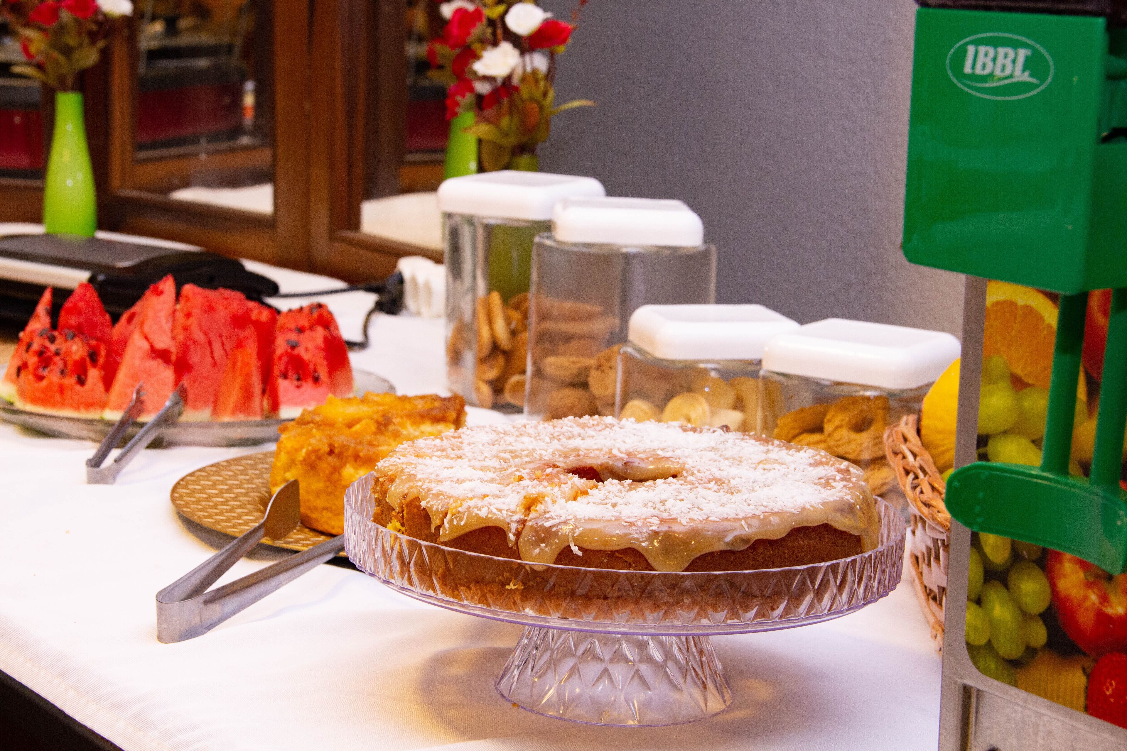 Una mesa de buffet con diversos alimentos, incluyendo un pastel sobre un pedestal de vidrio, una sandía y recipientes con nueces y frutas.
