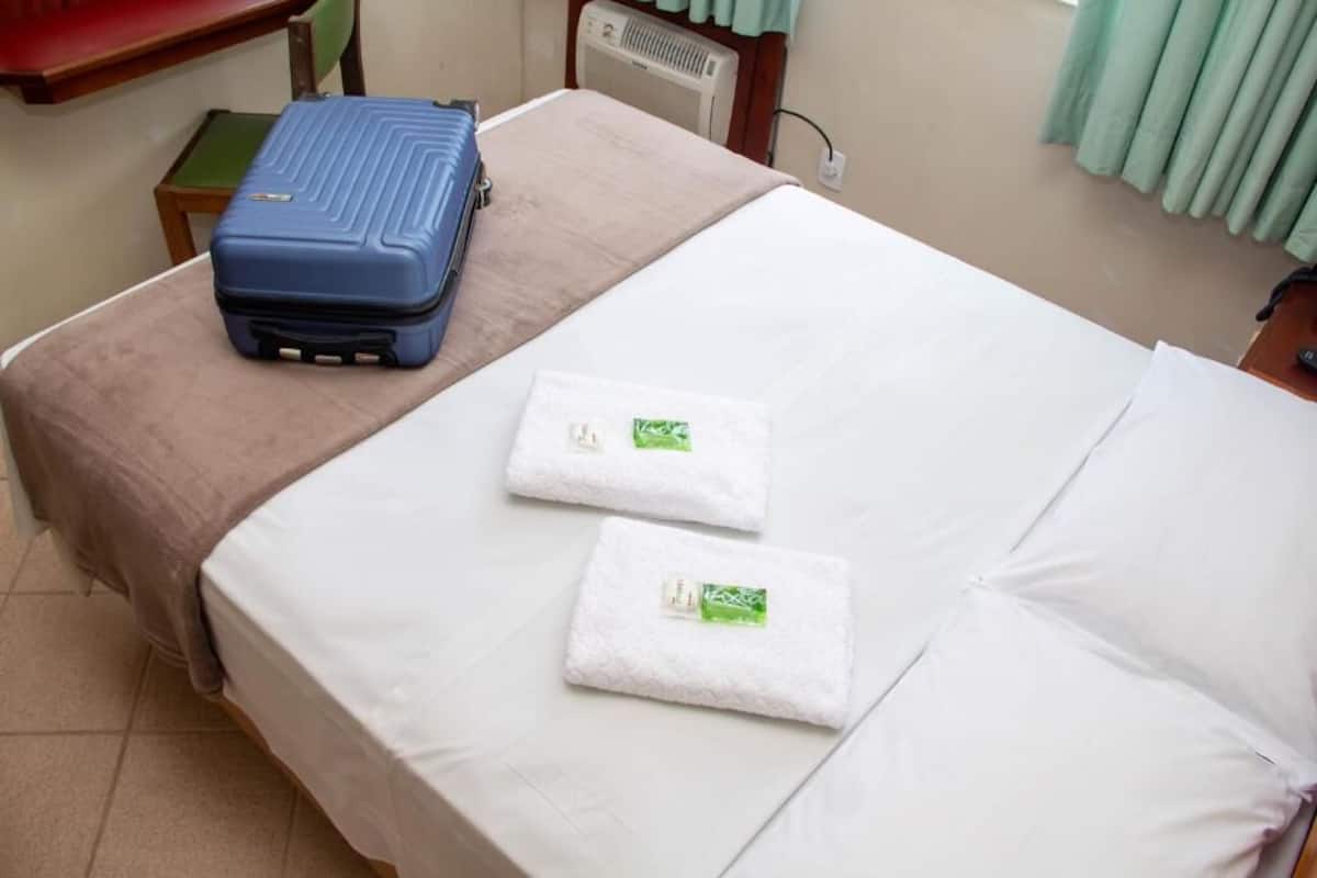 A neatly made bed with white linens and two folded towels with green tags. A blue suitcase is placed on a small table beside the bed.