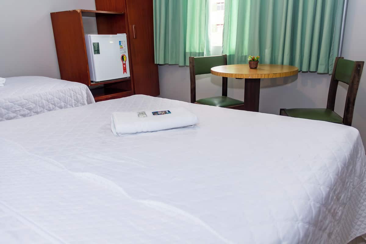 A hotel room with a bed, a small table, two chairs, a wooden wardrobe, and a refrigerator.