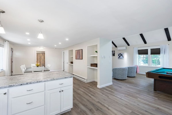 A modern kitchen with white cabinets, a marble countertop, and a pool table in a spacious living area with wooden flooring and built-in shelves.