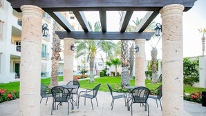 A covered outdoor seating area with a pergola, palm trees, and a view of a residential building in the background. - Condo PC by TRVL2HM (Cabo San Lucas)