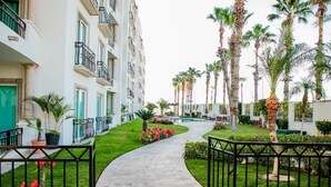 A pathway leading to a building with balconies and a garden with palm trees and flowers. - Condo PC by TRVL2HM (Cabo San Lucas)