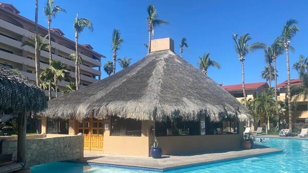 A pool area with a thatched-roof structure, palm trees, and a clear blue sky. - CONDO MRS BY TRVL2HM (Cabo San Lucas)