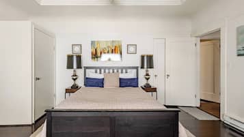 Quarto com uma cama grande, duas mesas de cabeceira com abajures, um quadro com uma vista da cidade na parede e uma porta que dá para outro cômodo.