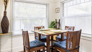 Uma área de refeições com mesa e cadeiras de madeira, um vaso com plantas secas e um quadro na parede.