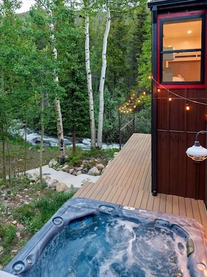 Un bain à remous situé dans un espace boisé, avec une terrasse en bois et un petit chalet illuminé de guirlandes lumineuses.