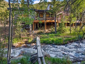 A cabin with a wooden deck, a river in front, and a forested backdrop.