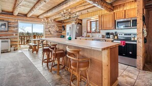 Cocina de cabaña de troncos con una isla central, taburetes de bar, armarios de madera y un ventanal grande.
