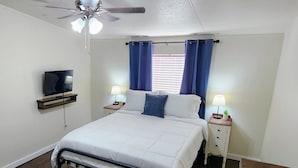 A bedroom with a bed, a ceiling fan, a TV on a shelf, two bedside tables with lamps, and a window with curtains.