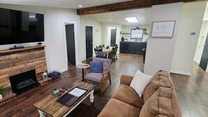 A living room with a fireplace, a sofa, a coffee table, and a TV. There is a kitchen in the background with a dining table and chairs.
