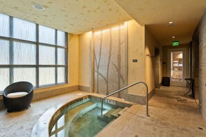 Een wellnessruimte met een hottub in glazen overkapping, een rieten stoel en een betegelde muur met een boommotief.