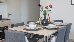 En matplats med ett bord dukat för fyra personer, omgivet av grå stolar. På bordet står två vaser med blommor, samt en flaska olivolja och en flaska apelsinjuice.