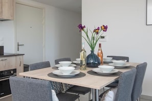 A dining area with a table set for four, surrounded by grey chairs. There are two vases with flowers on the table, and a bottle of olive oil and a bottle of orange juice.