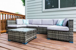 A sectional sofa with cushions, a glass-top coffee table, and wicker furniture on a wooden deck.