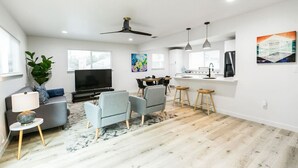 A modern living room with a flat-screen TV, grey sofas, a coffee table, and a dining area with bar stools. - EastSide Austin · Quaint 4 Bedroom Eastside Lodge (Pet Friendly & Work Space) (Austin)