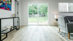 A living room with a sliding glass door to a backyard, a grey sofa, and a glass coffee table. - EastSide Austin · Quaint 4 Bedroom Eastside Lodge (Pet Friendly & Work Space) (Austin)