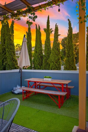 A patio with a wooden table and benches, surrounded by green artificial grass and tall trees. - ★Insta-Worthy Sunset View ★Private Rooftop ★Garage ★Silver Lake ★Dodgers Stadium (Los Angeles)