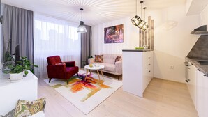 A modern living room with a red chair, a white sofa, a colorful rug, and a kitchen area with white cabinets and a stove.